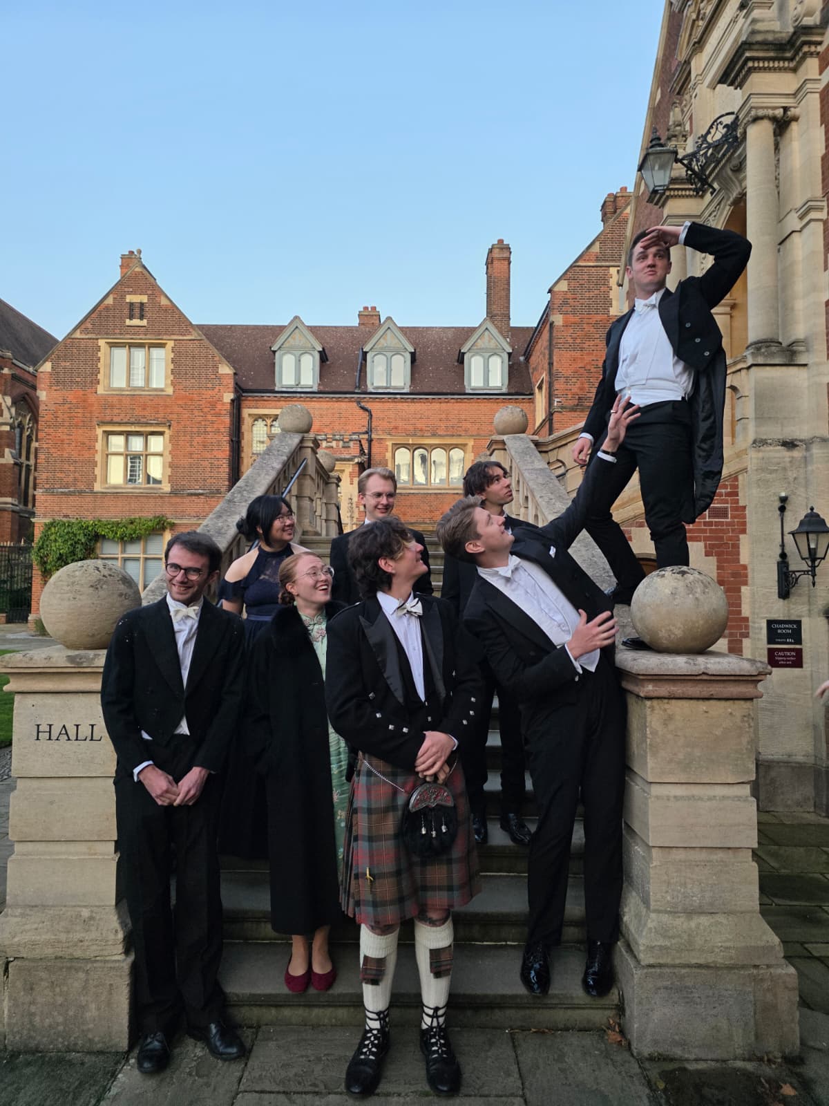 Members in white tie on the steps outside Hall, one standing on the balustrade above the others