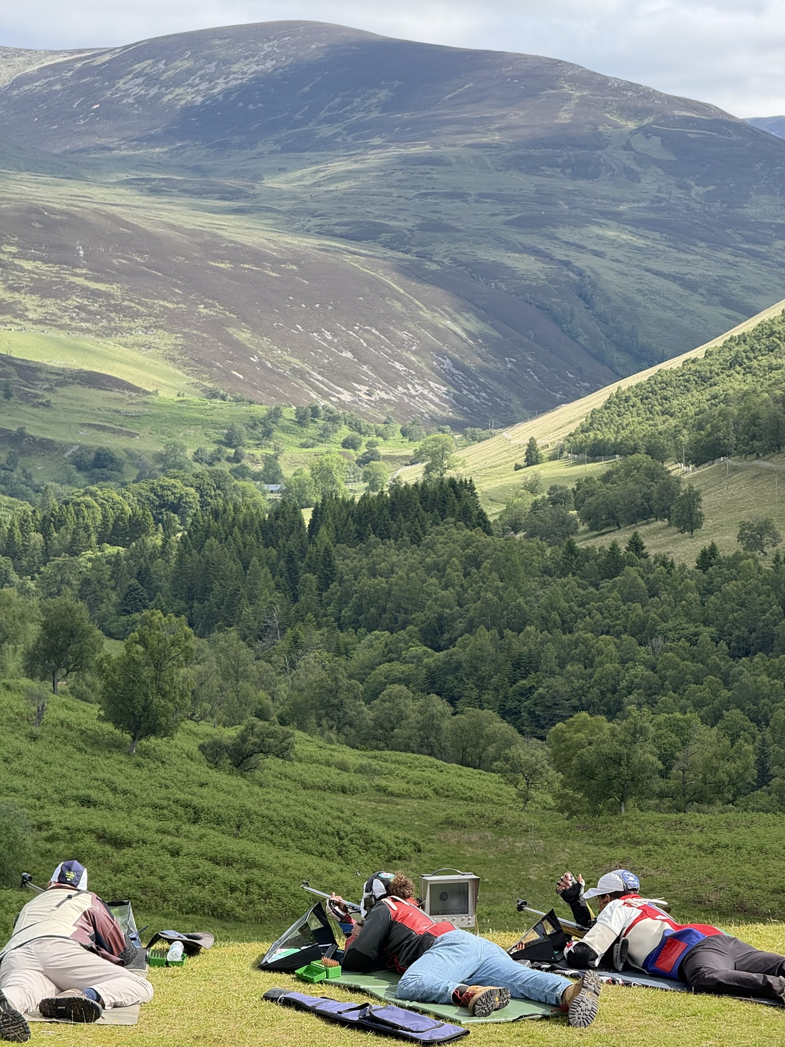Three shooters prone on the firing point at Blair Atholl, looking out over a forested glen
