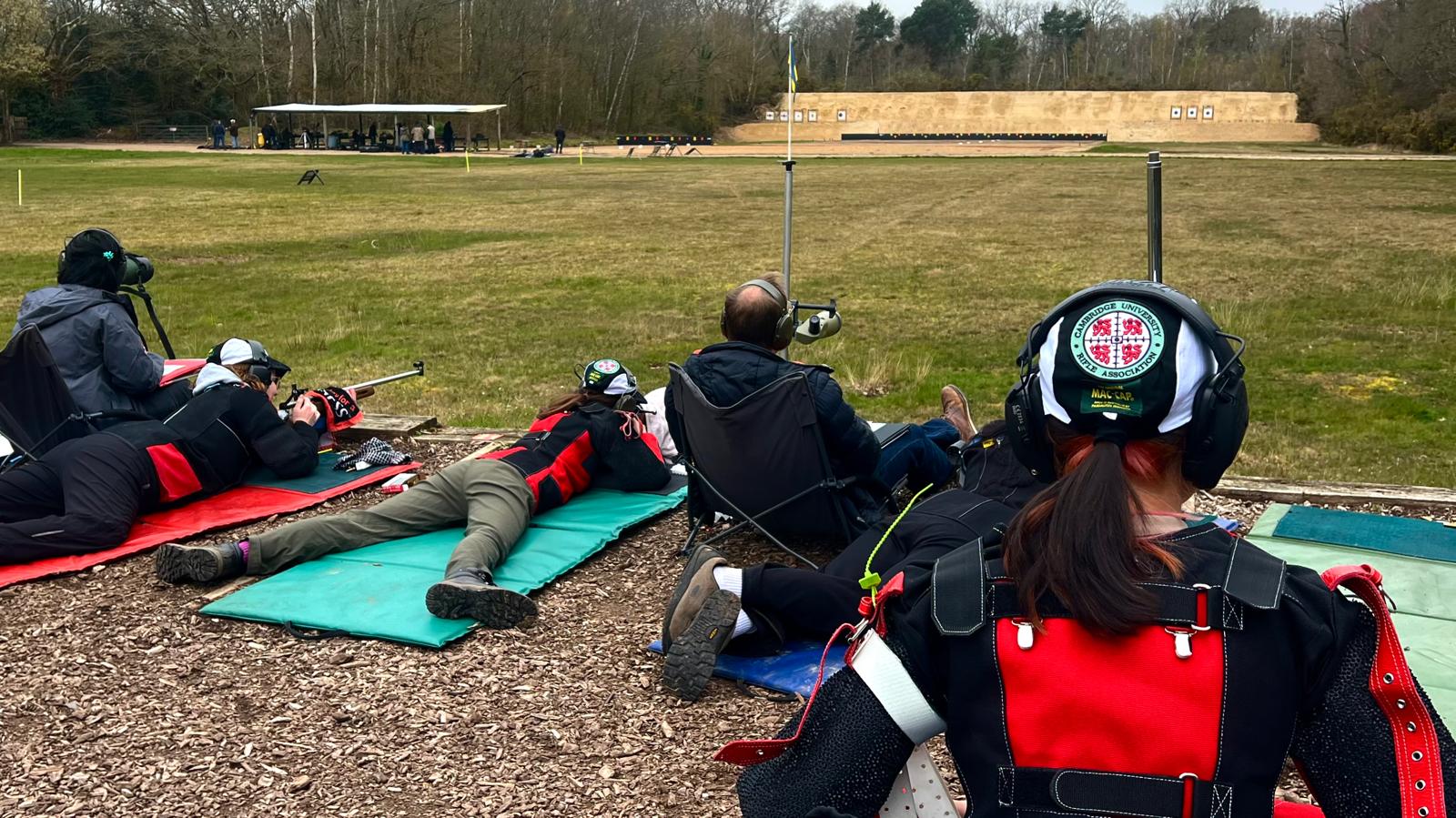 Shooters prone on mats at the firing point, a CURA ear-defender cap in the foreground