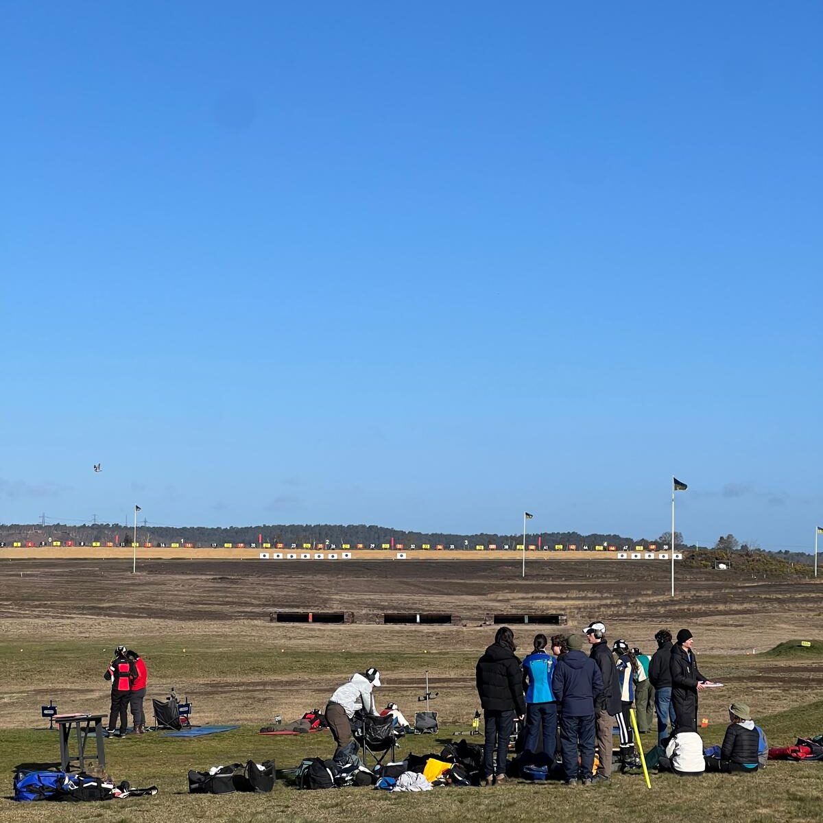 Members gathered with kit behind the firing point at Bisley under a clear sky, the target line beyond